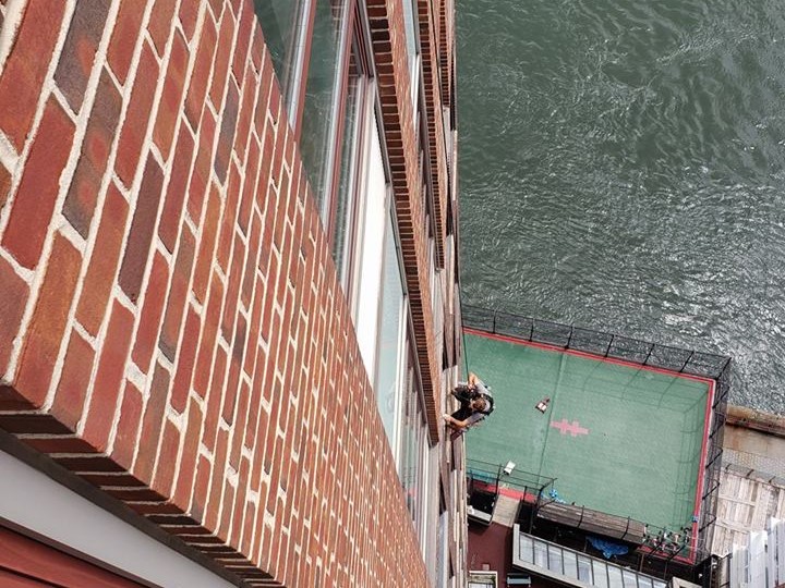 A focused rope access technician dangles deftly on vertical ropes against a high-rise building, equipped with professional cleaning tools, showcasing the precision and care taken in maintaining the sparkling facade of the urban landscape.