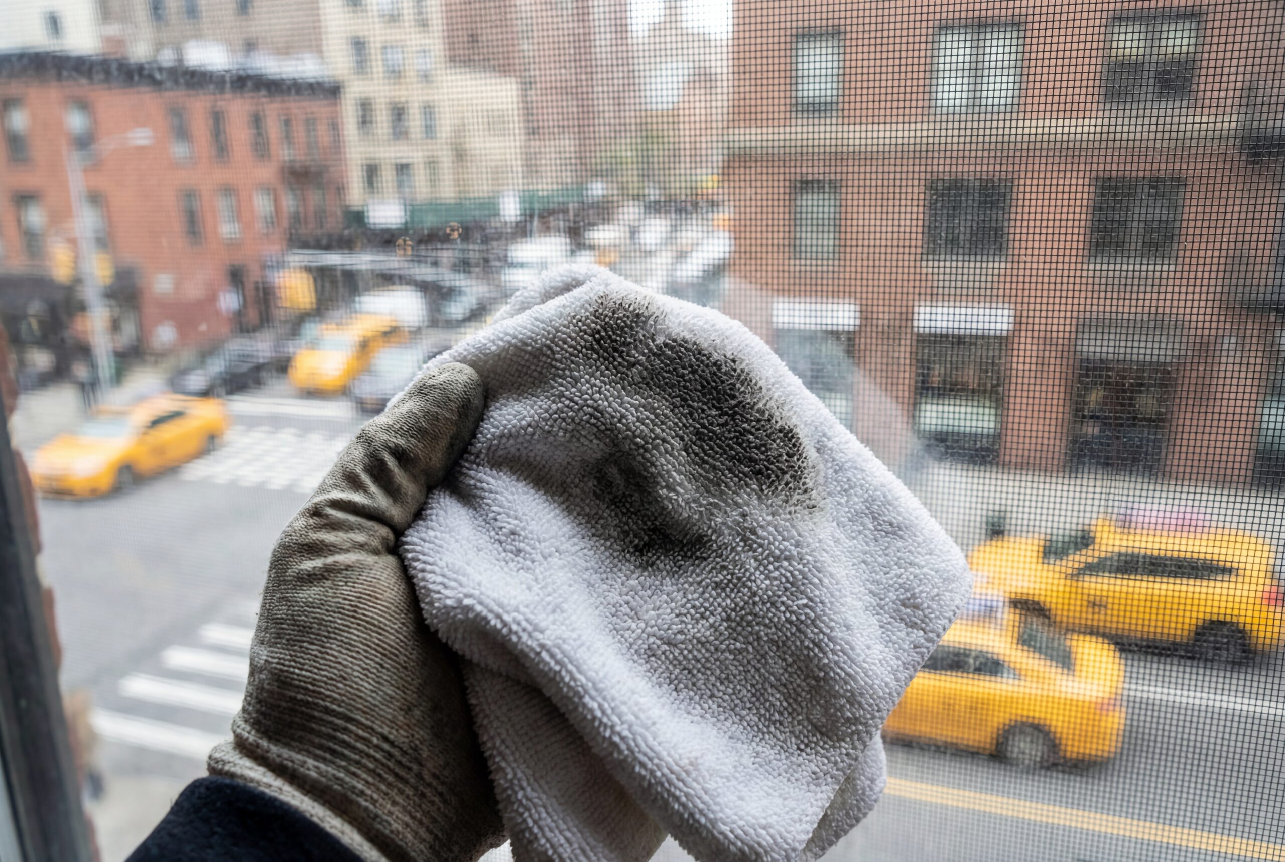 A hand holding a white microfiber cloth stained with black urban dust and soot after wiping a dirty NYC window screen.