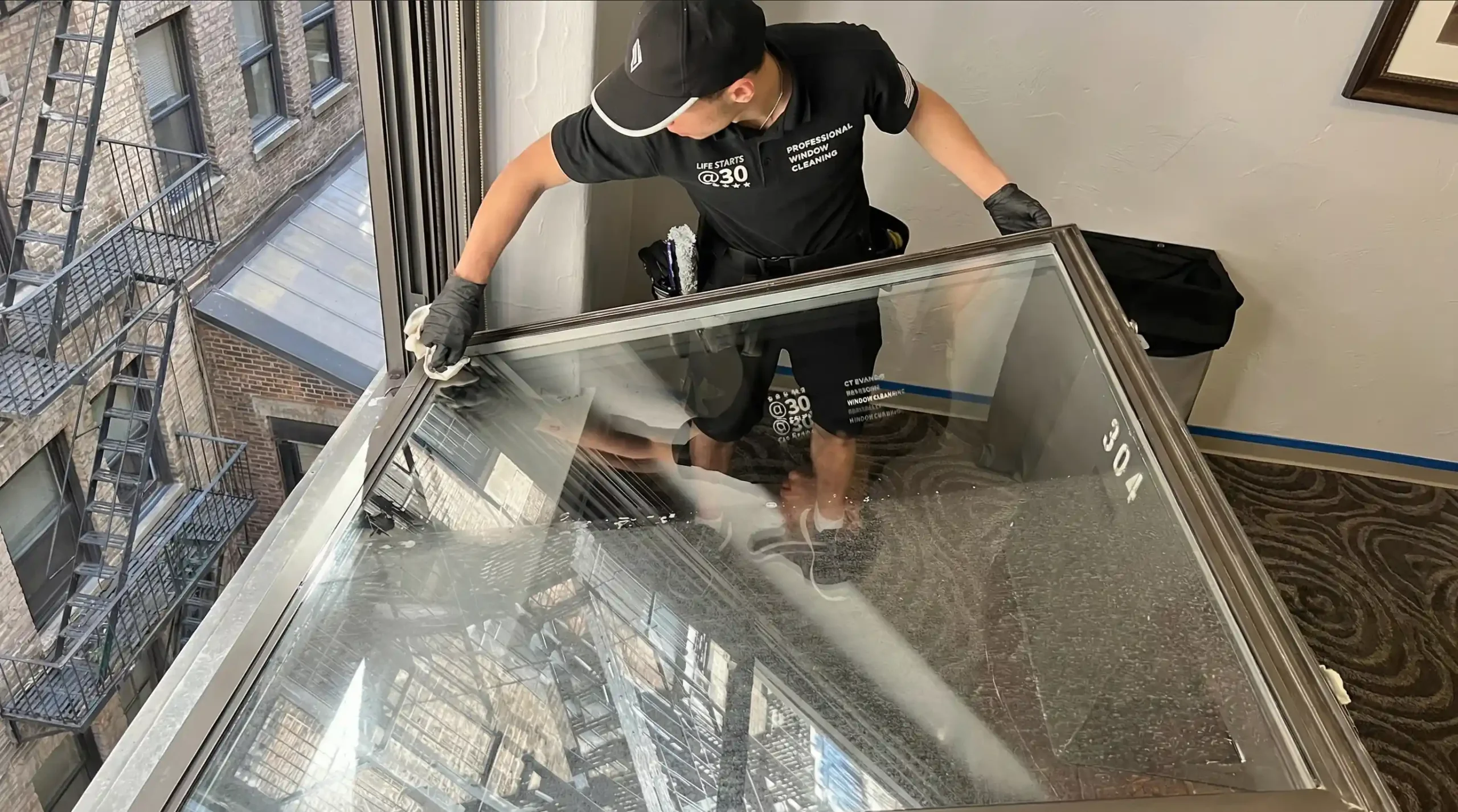 Big Apple Window Cleaning technician cleaning a tilt-in window from inside a Brooklyn apartment with a fire escape view