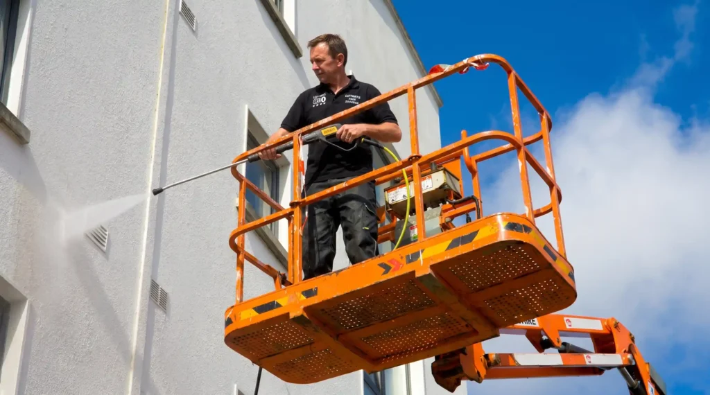 Big Apple Window Cleaning company technician on an orange boom lift using a pressure washer lance to clean a stucco commercial building facade