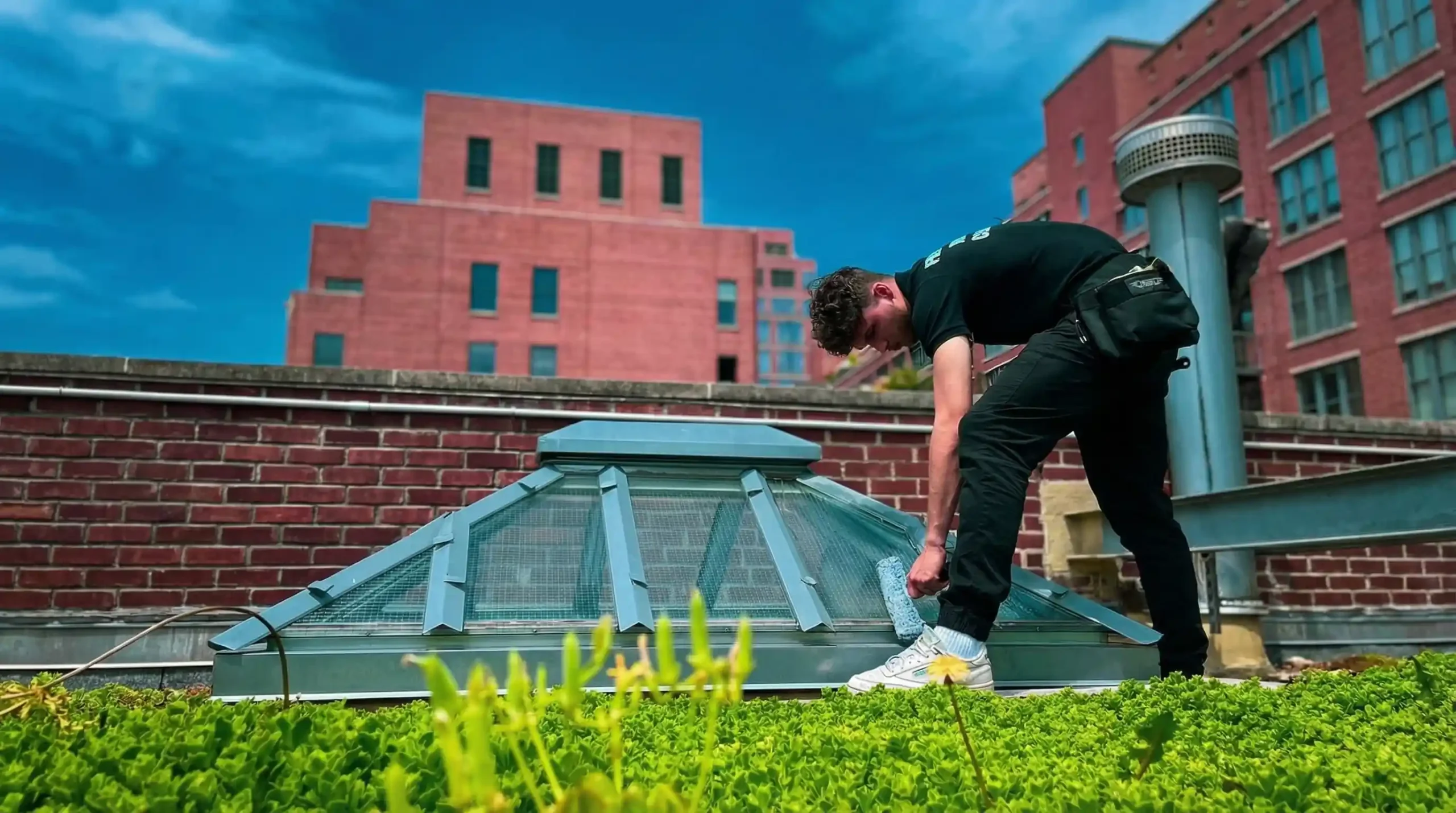 Big Apple Window Cleaning company technician cleaning a pyramid skylight on a green roof in New York City with brick buildings in the background