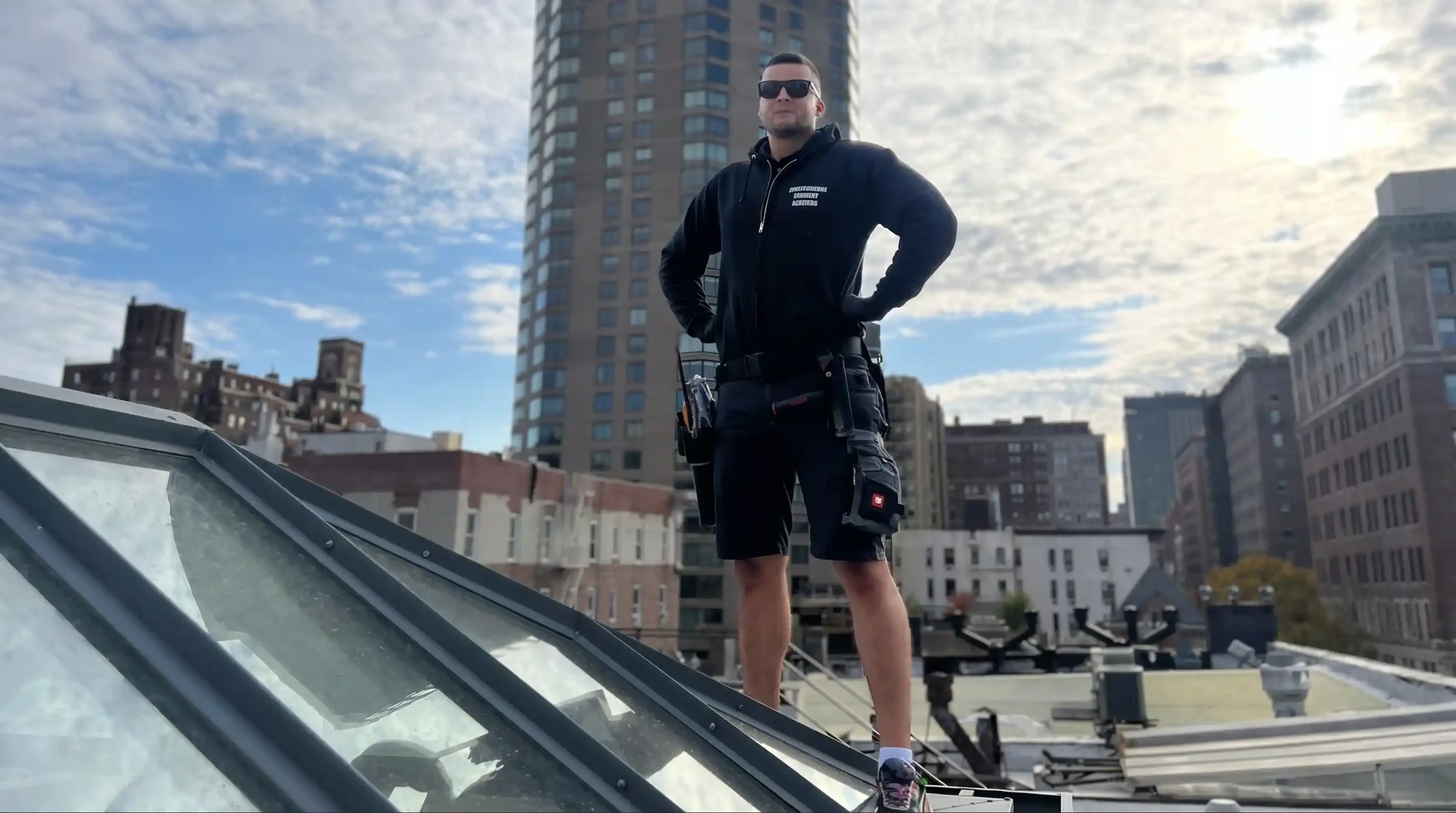A professional window cleaning technician (Big Apple Window Cleaning) stands on a New York City rooftop beside a glass roof lantern.
