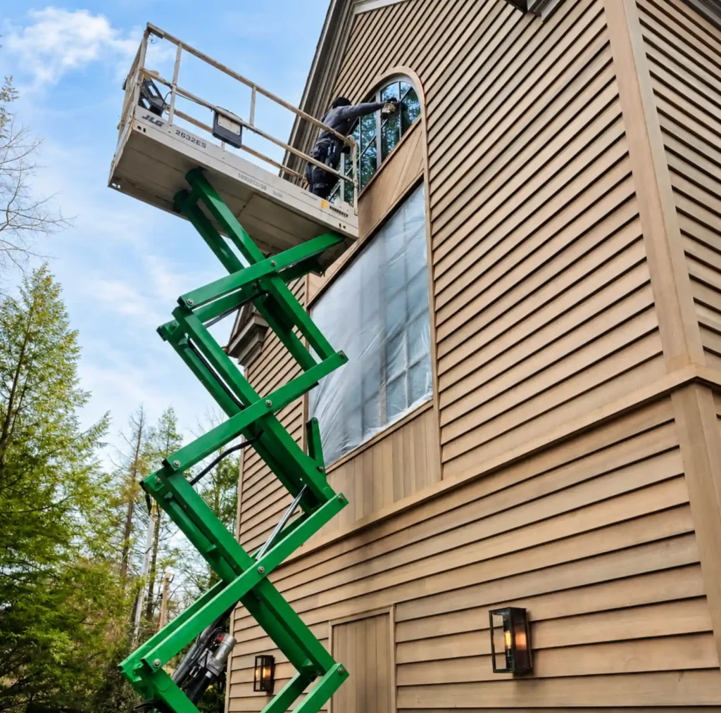 Technician on a JLG scissor lift removing protective film from a large arched window on a suburban New York home exterior