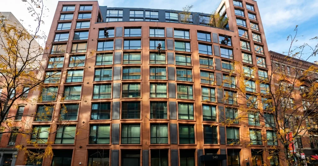 Three rope access window cleaners descending the brick and glass facade of a mid-rise Manhattan residential building in autumn
