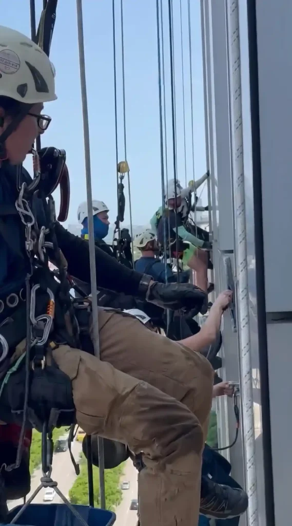 Group of rope access BAWC technicians in full harness and helmets working on a glass high-rise facade with street level visible below